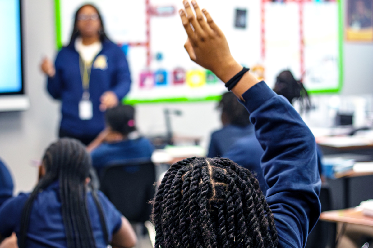 close up of student raising her hand with teacher blurred in the background